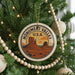 A circular ornament on a Christmas tree, featuring a desert landscape with mountains and a monument, is hanging from a string of beads.