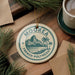 A circular ornament with the text "MOORE FRENCH POLYNESIA" printed on it, surrounded by pine branches and a cup of coffee on a wooden surface.