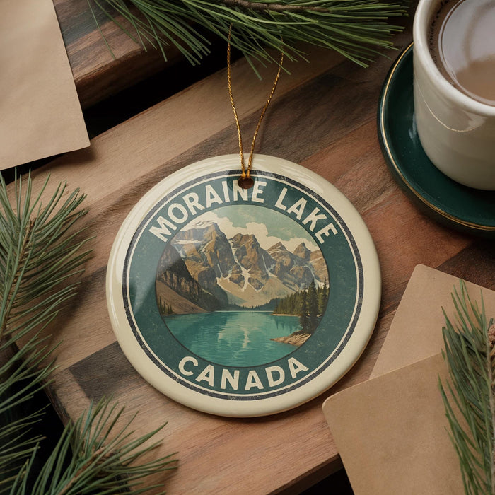 A circular ornament with the text "MORaine Lake Canada" and a scenic mountain landscape is displayed on a wooden surface, accompanied by pine branches and a cup of coffee.