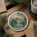 A circular ornament with the text "MORaine Lake Canada" and a scenic mountain landscape is displayed on a wooden surface, accompanied by pine branches and a cup of coffee.