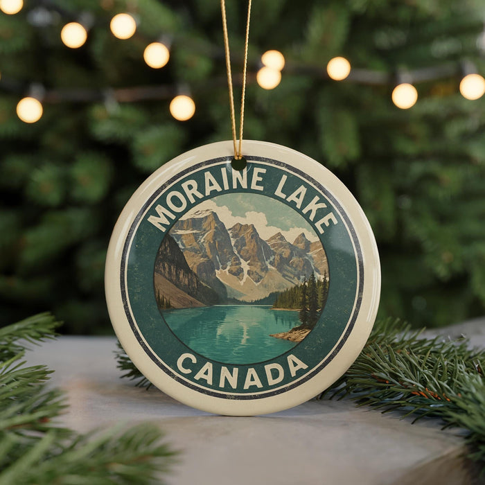 A circular ornament with a mountain landscape and a lake, surrounded by greenery, is displayed in front of a blurred background with Christmas lights.