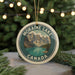 A circular ornament with a mountain landscape and a lake, surrounded by greenery, is displayed in front of a blurred background with Christmas lights.