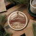 A circular ornament with a mountain landscape and the text "Mount Cook New Zealand" is displayed on a wooden surface, surrounded by pine branches and a cup of coffee.