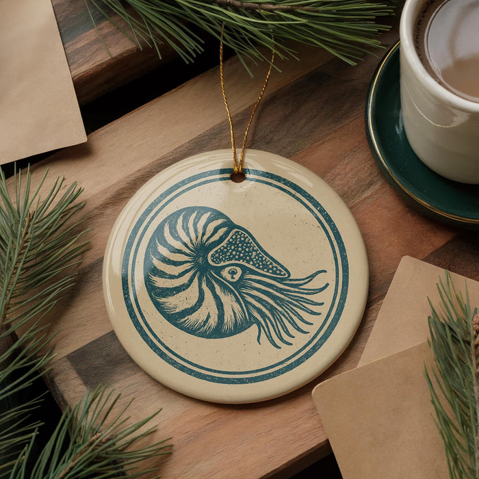 a round ornament with a blue and white design, resembling a nautilus shell, placed on a wooden surface. There is also a cup of coffee and some pine branches in the background.