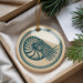 A round ornament with a blue and white design featuring a nautilus shell is displayed in a box, surrounded by pine branches.