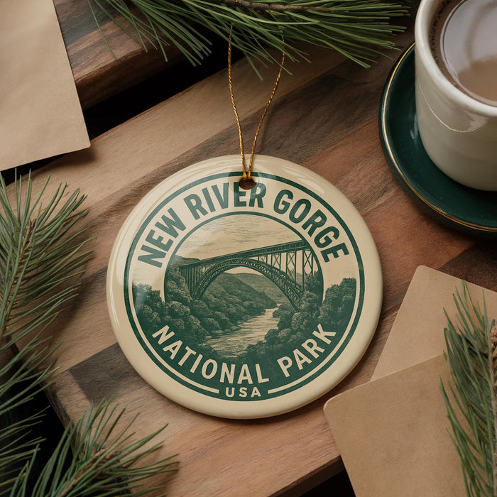 A circular ornament with the text "NEW RIVER GORGE NATIONAL PARK USA" and an illustration of a bridge, placed on a wooden surface alongside a cup of coffee and some pine branches.