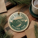 A circular ornament with the text "NEW RIVER GORGE NATIONAL PARK USA" and an illustration of a bridge, placed on a wooden surface alongside a cup of coffee and some pine branches.