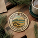 A decorative ornament with the text "NGORONGORO TANZANIA" and a landscape illustration, placed on a wooden surface next to a cup of coffee and pine branches.