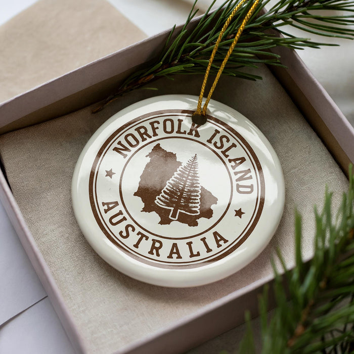 A white ornament with brown text and a circular design, featuring a tree silhouette, is displayed in a box with pine branches.