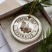 A white ornament with brown text and a circular design, featuring a tree silhouette, is displayed in a box with pine branches.