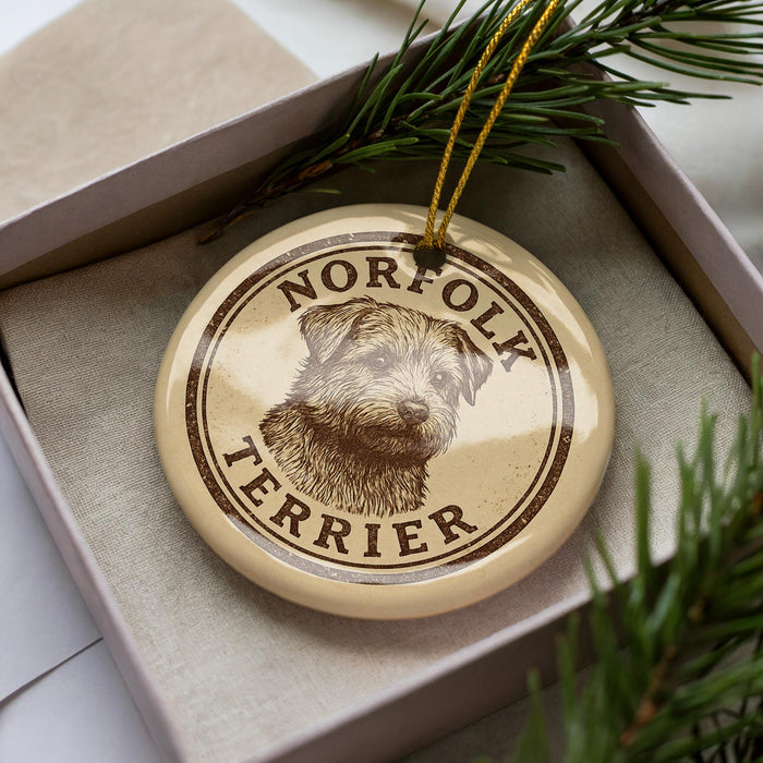 A round ornament with a dog's head and the text "NORFOLK TERRIER" printed on it, placed inside a box with pine branches.