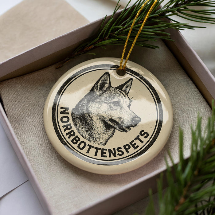 A circular ornament with a black and white illustration of a dog's head and the text "NORBOTTTENSPEPS" on it, placed inside a box with pine branches.