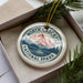 A decorative ornament in a box, featuring a mountain landscape with pine trees and a river, along with the text "NORTH CASCADES NATIONAL PARK USA".