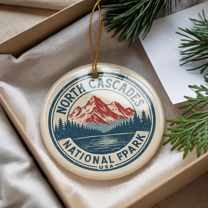 A decorative ornament with a mountain and pine tree design, featuring the text "NORTH CASCADES NATIONAL FPLAY" on it, is displayed in a box with pine branches.