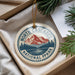 A decorative ornament with a mountain and pine tree design, featuring the text "NORTH CASCADES NATIONAL FPLAY" on it, is displayed in a box with pine branches.
