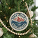 A Christmas tree decorated with ornaments, including a circular ornament with a mountain and lake design, surrounded by lights and greenery.