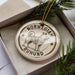 A round ornament with a dog image and the text "NORWEGIAN Buhund" on it, placed in a box with pine branches.