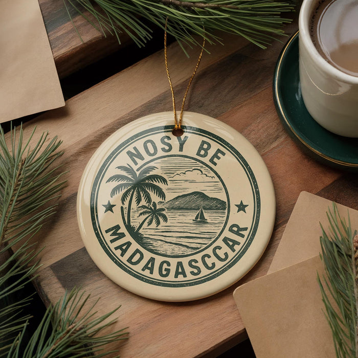 A circular ornament with the text "NOSY BE MADAGASCAR" and a palm tree illustration, placed on a wooden surface next to a cup of coffee and some pine branches.