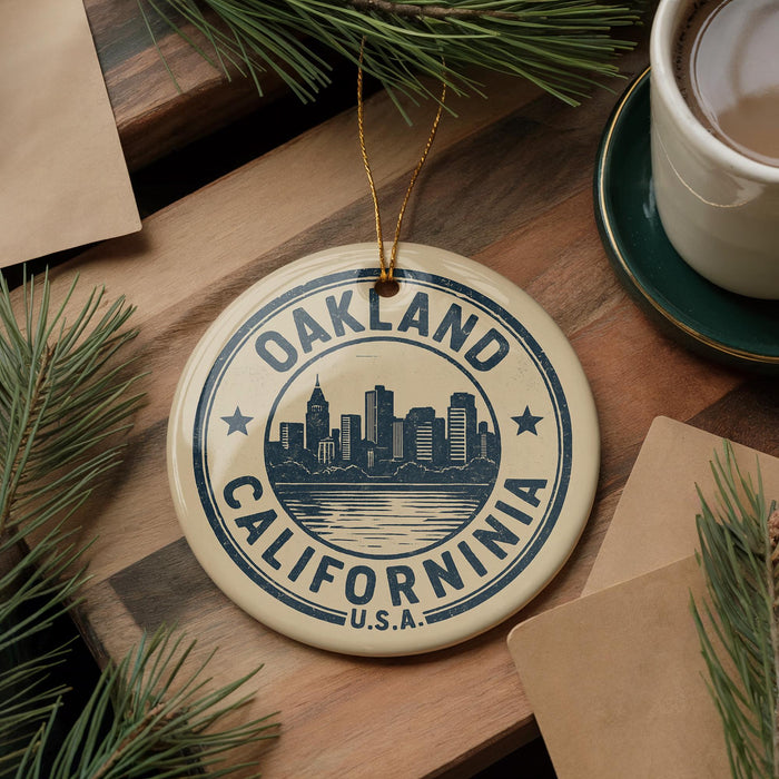 A circular ornament with the text "OAKLAND CALIFORNIA" and a graphic of a city skyline, hanging on a string. The ornament is placed on a wooden surface, surrounded by pine branches and a cup of coffee.