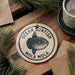 A round ornament featuring a fish, likely an ocean sunfish, is displayed on a wooden surface alongside a cup of coffee and some pine branches.