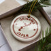 A white ornament with a red border and the text "OKINAWA JAPAN" is displayed in a box, surrounded by pine branches.