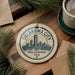 A circular ornament with the text "OKLAHOMA CITY USA" and a graphic of a city skyline is displayed on a wooden surface, surrounded by pine branches and a cup of coffee.