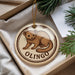 A round ornament with a brown animal, possibly a rodent or small mammal, on it, placed in a box with pine branches.