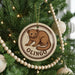 A round ornament with a brown bear image and the word "OLINGO" is hanging on a Christmas tree.