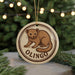A round ornament featuring a brown cartoon-like animal, possibly a bear or a similar creature, with the word "OLINGO" written around it. The ornament is hanging from a string, and there are pine branches visible in the background.