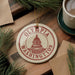 A circular ornament with the text "OLYMPIA WASHINGTON" and a graphic of the Washington State Capitol building, placed on a wooden surface alongside pine branches and a cup of coffee.