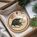 A round ornament with the word "OPOSSIUM" printed on it, featuring an illustration of a small animal, is displayed in a box with pine branches.