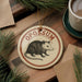 A circular ornament with the word "OPOSSIUM" printed on it, featuring an illustration of a small opossum. The ornament is hanging from a string, and there are pine branches and a cup of coffee visible in the background.