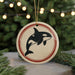 A circular ornament featuring a black and white orca whale silhouette is displayed against a blurred background of Christmas lights and pine branches.