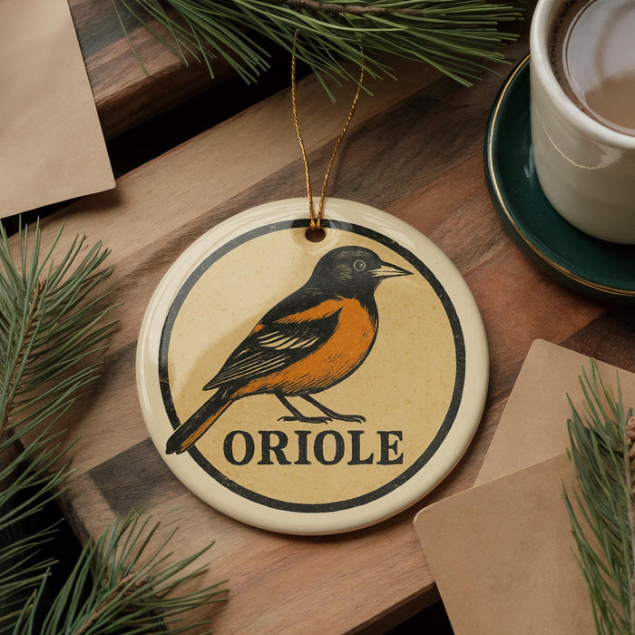 A ceramic ornament featuring an illustration of an oriole bird, with the word "ORIOLLE" written below it, is displayed on a wooden surface alongside a cup of coffee and some pine branches.