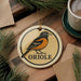 A ceramic ornament featuring an illustration of an oriole bird, with the word "ORIOLLE" written below it, is displayed on a wooden surface alongside a cup of coffee and some pine branches.