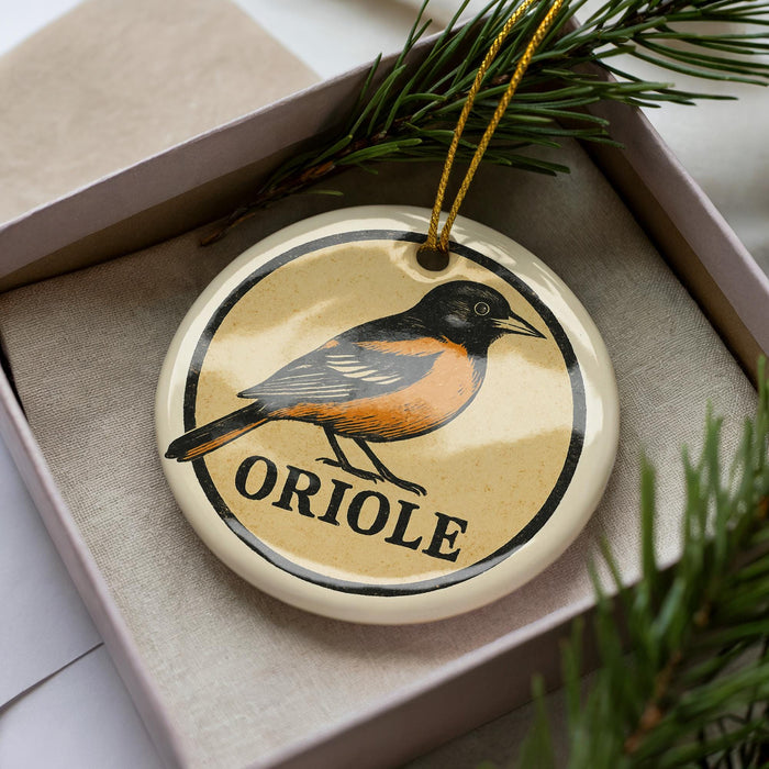 A ceramic ornament in the shape of a bird, specifically an oriole, is displayed in a box with pine branches.