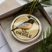 A ceramic ornament in the shape of a bird, specifically an oriole, is displayed in a box with pine branches.