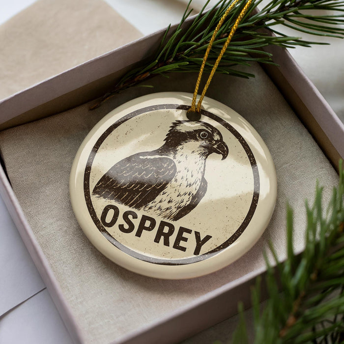 A circular ornament with a bird image and the word "OSPREY" printed on it, placed inside a box with pine branches.