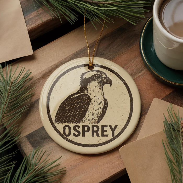 A ceramic ornament featuring an illustration of an Osprey bird, with the word "OSPREY" written on it, is displayed on a wooden surface alongside a cup of coffee and some pine branches.