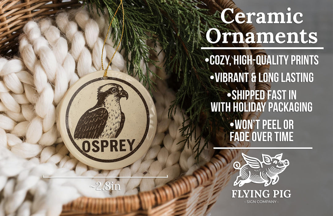 Ceramic ornaments, featuring a ceramic ornament with an Osprey image and text, placed in a wicker basket with pine branches.