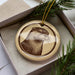 A ceramic ornament featuring an image of an otter is displayed in a box, surrounded by pine branches.
