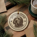 A circular ornament featuring a dog's head and the word "OTTERHOUND" is displayed on a wooden surface, surrounded by pine branches and a cup of coffee.