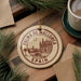 A circular ornament with the text "PALMA DE MALLORCA SPAIN" printed on it, surrounded by pine branches and a cup of coffee on a wooden surface.
