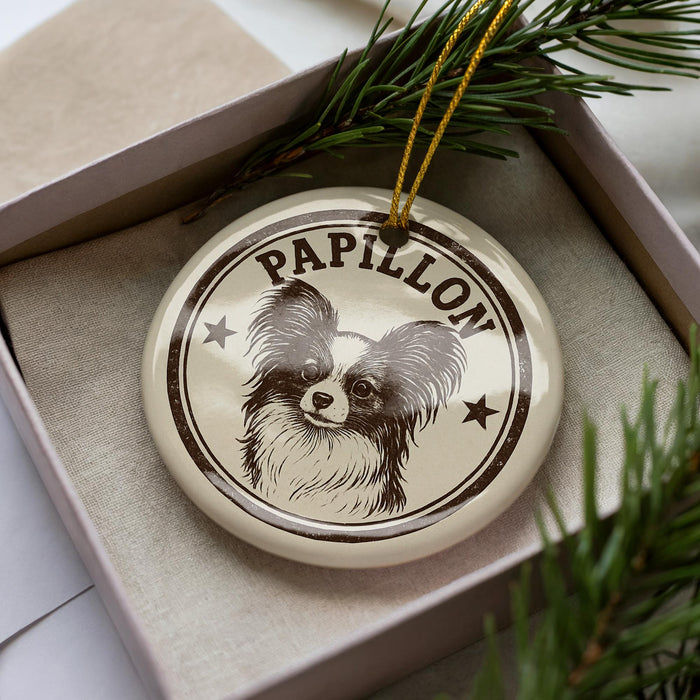 A decorative ornament featuring a Papillon dog, with the name "Papillon" written on it, is displayed in a box surrounded by pine branches.