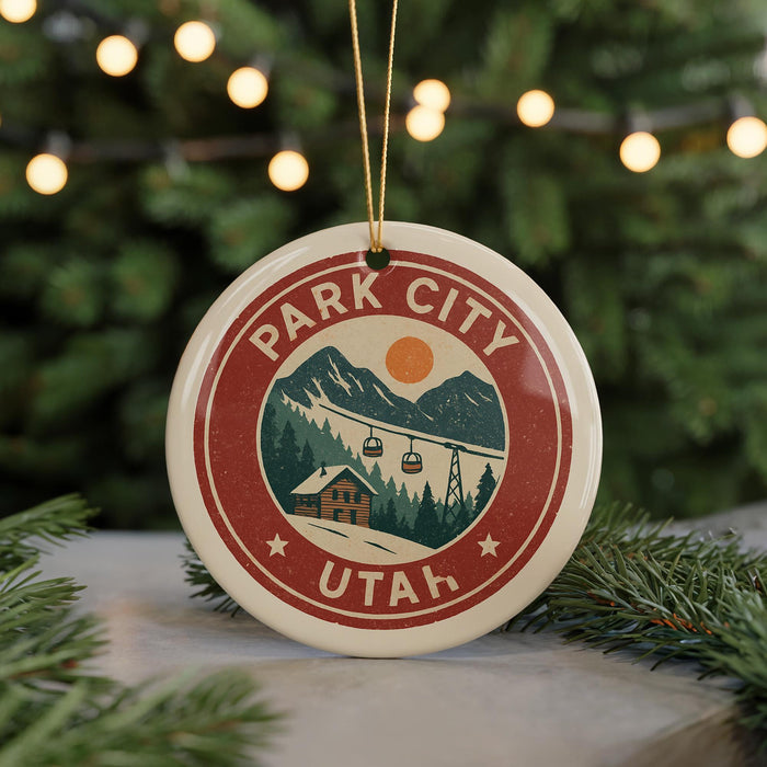 A circular ornament with the text "PARK CITY UTAH" and an illustration of a mountain landscape with ski lifts, hanging from a string against a blurred background of Christmas lights.