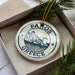 A circular ornament with the text "PAROS GREECE" and an illustration of a Greek island, placed in a box with pine branches.