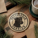 A circular ornament featuring a black dog's head, with the text "PATTERDALE TERRIER" around the edge, is displayed on a wooden surface alongside a cup of coffee and some pine branches.
