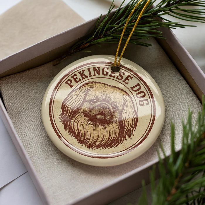 A circular ornament with a picture of a dog's face and the text "Pekingese Dog" on it, placed in a box with pine branches.