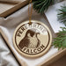 A round ornament with the text "Peregrine Falcon" and an illustration of a peregrine falcon is displayed in a box with pine branches.