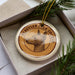 A round ornament with a dog's head on it, placed inside a box with pine branches.